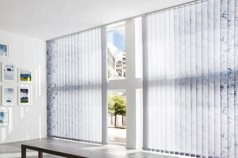 Modernes Zimmer mit großen vertikalen Jalousien, die teilweise geöffnet sind und das Sonnenlicht hereinlassen. Vor dem Fenster steht ein einfacher Tisch, und an der angrenzenden weißen Wand hängen gerahmte Kunstwerke. Draußen sind Bäume und ein Gebäude zu sehen.