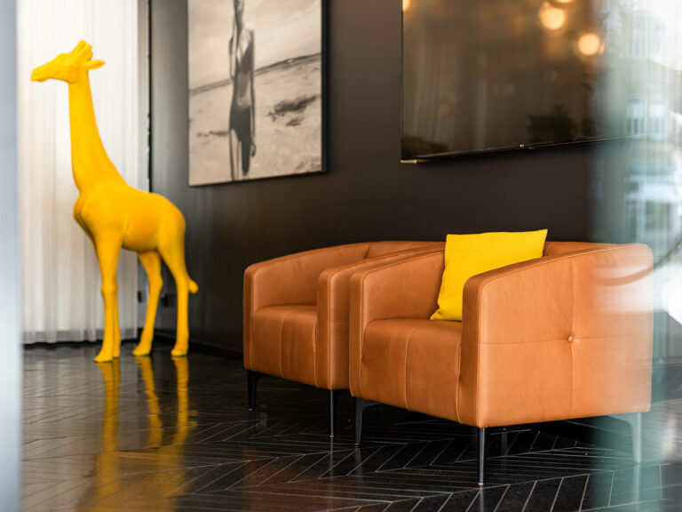 Zwei hellbraune Sessel mit gelben Kissen stehen neben einer großen gelben Giraffenskulptur in einem modernen Empfangsbereich mit dunklen Wänden, einem Schwarz-Weiß-Foto und einem Fernsehbildschirm.