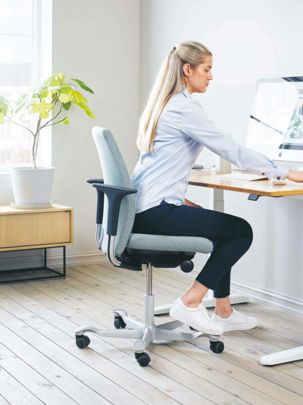 Eine Frau mit langen blonden Haaren sitzt auf einem ergonomischen Bürostuhl an einem modernen Schreibtisch und konzentriert sich auf ihren Computer. Der Arbeitsplatz ist mit Details der Büroeinrichtung ausgestattet: helle Holzböden, reichlich natürliches Licht, eine Pflanze und eine grüne Tasche auf dem Boden.