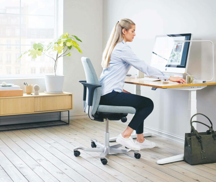 Eine Frau mit langen blonden Haaren sitzt auf einem ergonomischen Bürostuhl an einem modernen Schreibtisch und konzentriert sich auf ihren Computer. Der Arbeitsplatz ist mit Details der Büroeinrichtung ausgestattet: helle Holzböden, reichlich natürliches Licht, eine Pflanze und eine grüne Tasche auf dem Boden.