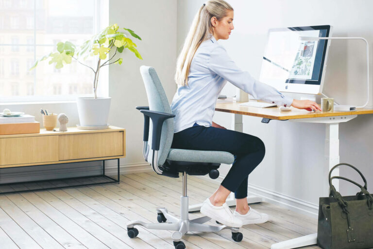 Eine Frau mit langen blonden Haaren sitzt auf einem ergonomischen Bürostuhl an einem modernen Schreibtisch und konzentriert sich auf ihren Computer. Der Arbeitsplatz ist mit Details der Büroeinrichtung ausgestattet: helle Holzböden, reichlich natürliches Licht, eine Pflanze und eine grüne Tasche auf dem Boden.