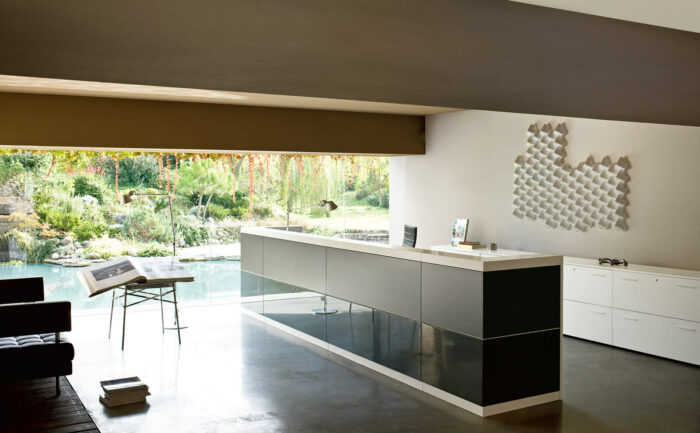 Modernes, minimalistisches Büro mit einem langen, schlanken Schreibtisch vor einem großen Fenster mit Blick auf einen üppigen Garten und einen Teich. Perfekt für den Empfang oder konzentriertes Arbeiten, mit offenen Büchern auf einem Beistelltisch, weißen Schränken und geometrischer Wandkunst.