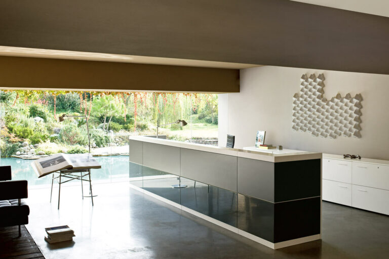 Frezza_Vogue Modernes, minimalistisches Büro mit einem langen, schlanken Schreibtisch vor einem großen Fenster mit Blick auf einen üppigen Garten und einen Teich. Perfekt für den Empfang oder konzentriertes Arbeiten, mit offenen Büchern auf einem Beistelltisch, weißen Schränken und geometrischer Wandkunst.