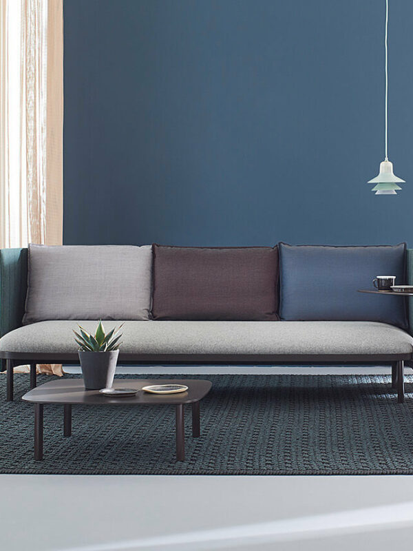 Ein modernes Wohnzimmer mit einer blauen Wand, hellen Vorhängen, einem grauen Sofa mit verschiedenen Kissen, zwei Sesseln, einem Couchtisch, einer Stehlampe, Pflanzen und einer Hängelampe auf einem gemusterten Teppich.