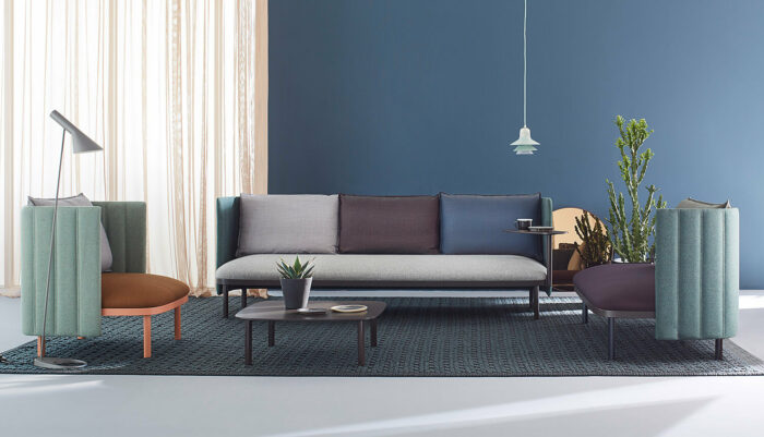 Ein modernes Wohnzimmer mit einer blauen Wand, hellen Vorhängen, einem grauen Sofa mit verschiedenen Kissen, zwei Sesseln, einem Couchtisch, einer Stehlampe, Pflanzen und einer Hängelampe auf einem gemusterten Teppich.