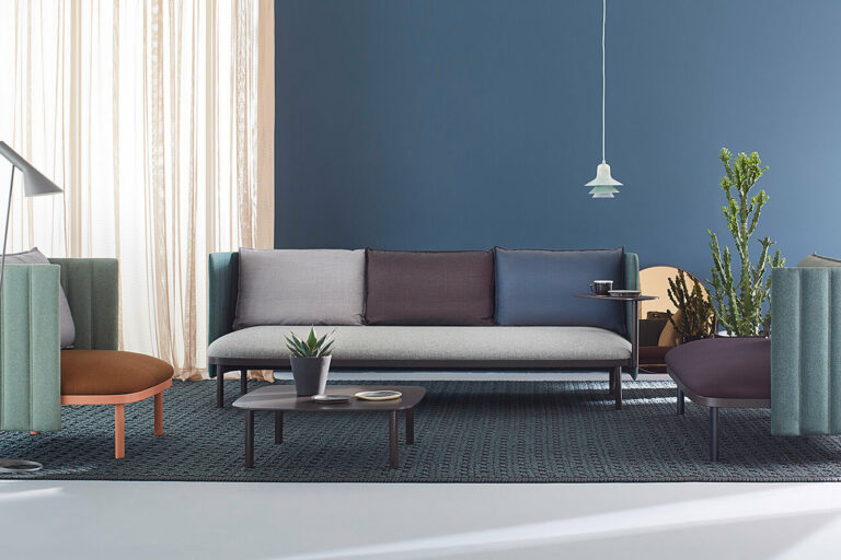 knezevic_lounge_ophelis_sum_3er_Konfiguration_Paravent_niedrig Ein modernes Wohnzimmer mit einer blauen Wand, hellen Vorhängen, einem grauen Sofa mit verschiedenen Kissen, zwei Sesseln, einem Couchtisch, einer Stehlampe, Pflanzen und einer Hängelampe auf einem gemusterten Teppich.