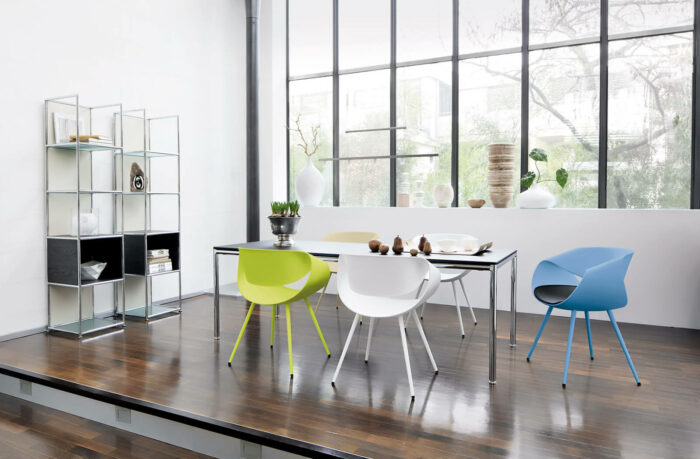 Moderner Essbereich mit dunklem Holzfußboden, einem Glastisch, bunten Stühlen (weiß, grün, blau) und einem Metallregal mit Dekorationsartikeln, das an stilvolle Konferenzräume erinnert, vor großen Fenstern mit Blick auf Bäume.