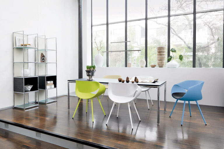 tw_Essen_01_buntweb2 Moderner Essbereich mit dunklem Holzfußboden, einem Glastisch, bunten Stühlen (weiß, grün, blau) und einem Metallregal mit Dekorationsartikeln, das an stilvolle Konferenzräume erinnert, vor großen Fenstern mit Blick auf Bäume.