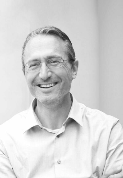 Portrait von Thomas Knezevic, der ein helles Button-up-Hemd trägt und vor einem schlichten, hellen Hintergrund steht, lächelt freundlich. Das Foto ist in schwarz-weiß gehalten.