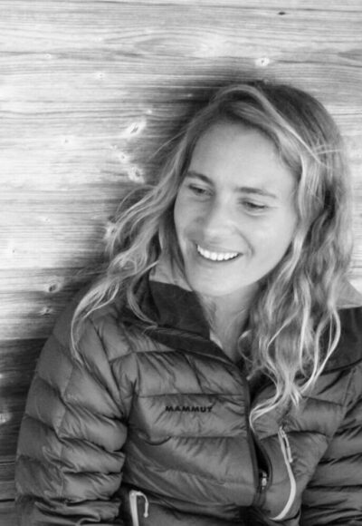 Portrait von Sandra Aumeier sitzt da und lächelt, wobei sie leicht zur Seite schaut. Der Hintergrund ist eine Holzvertäfelung. Das Bild ist in schwarz-weiß gehalten.